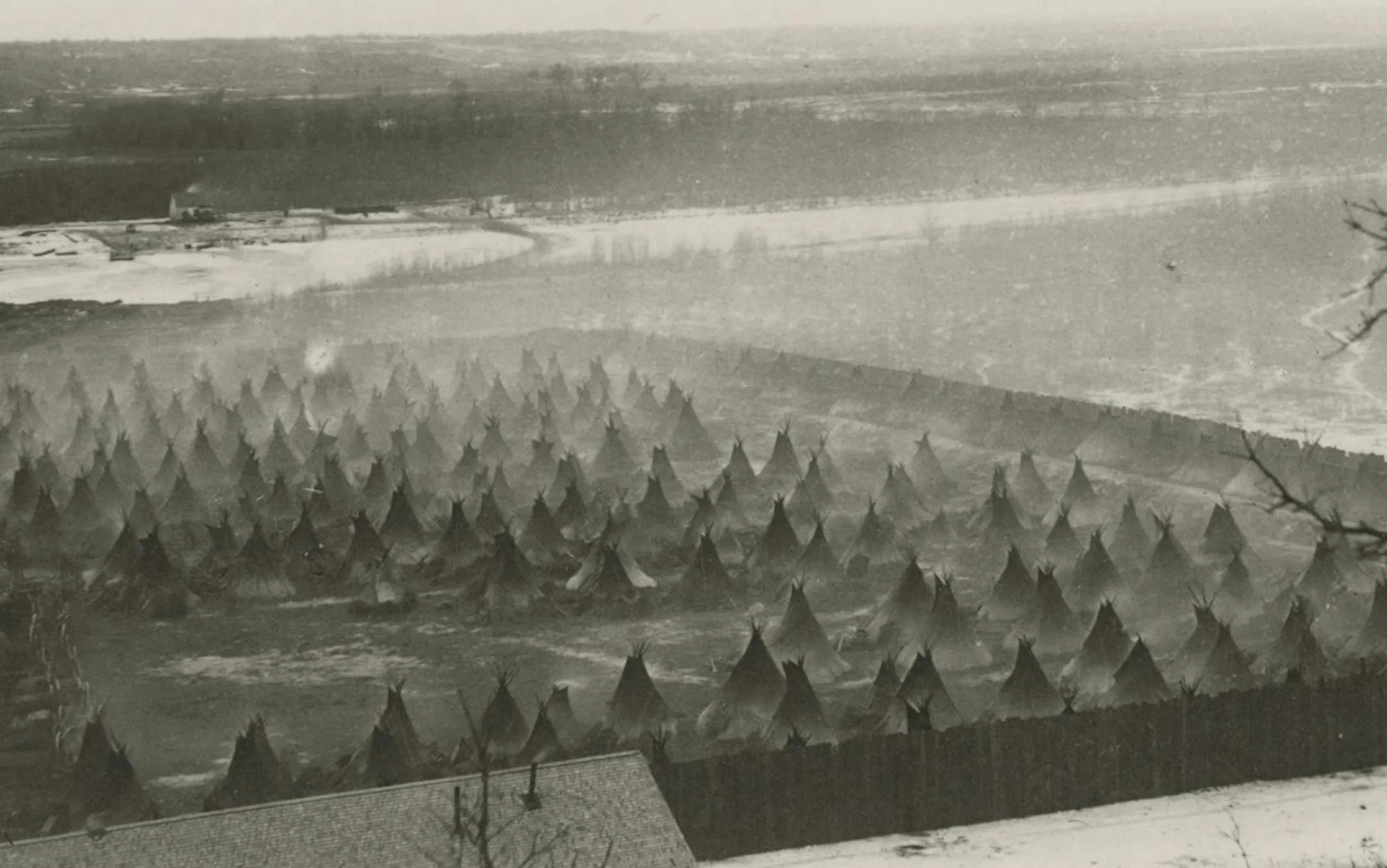 A photograph shows dozens of tepees fenced in by a tall stockade. The ground appears to be frozen mud. Smoke rises from several tepees, the result of warming fires within