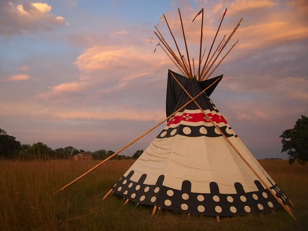 A colorful tepee stands in tall prairie grass at sunset, with a stone Agency building in the far background